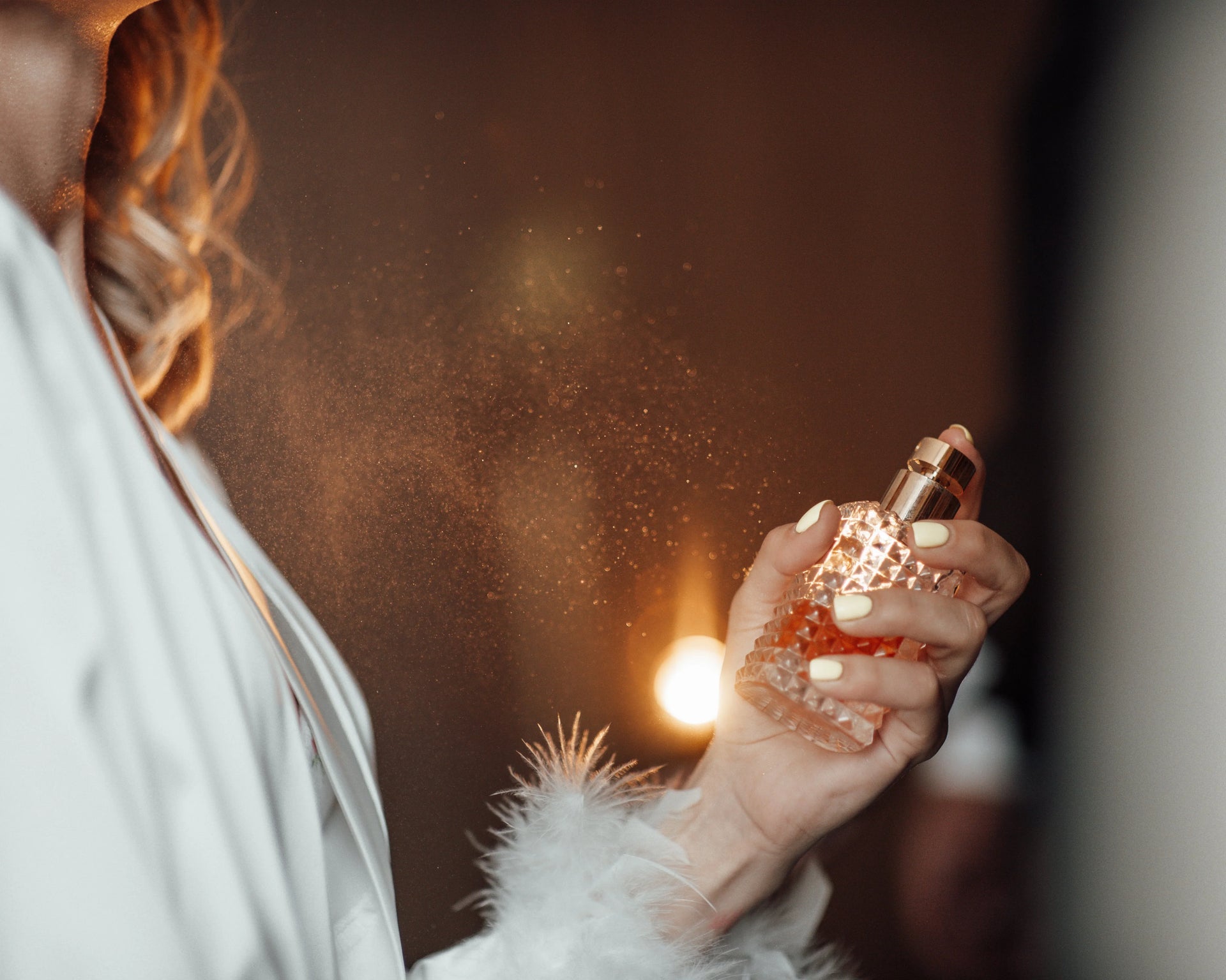 Woman spraying unisex perfume from a textured glass bottle in a soft, elegant setting.