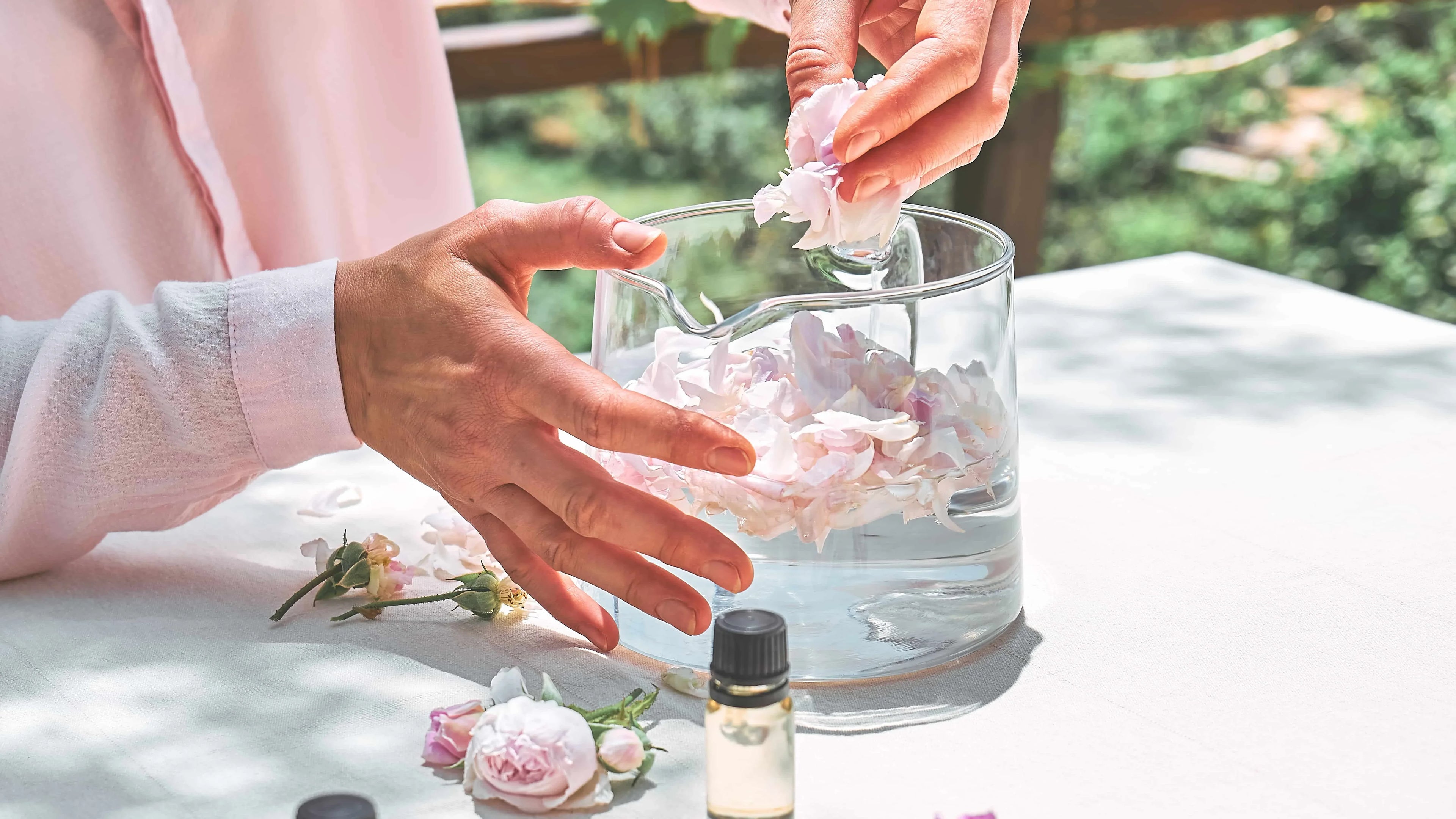 Woman placing fresh flower petals in glass container for natural perfume making outdoors